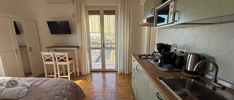 A compact hotel room with a bed, a small dining area, a kitchenette, and a view through a glass door.