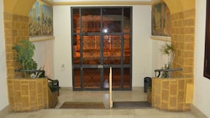 Entrada de hotel con un amplio ventanal de vidrio, pilares de piedra y suelo de baldosas.