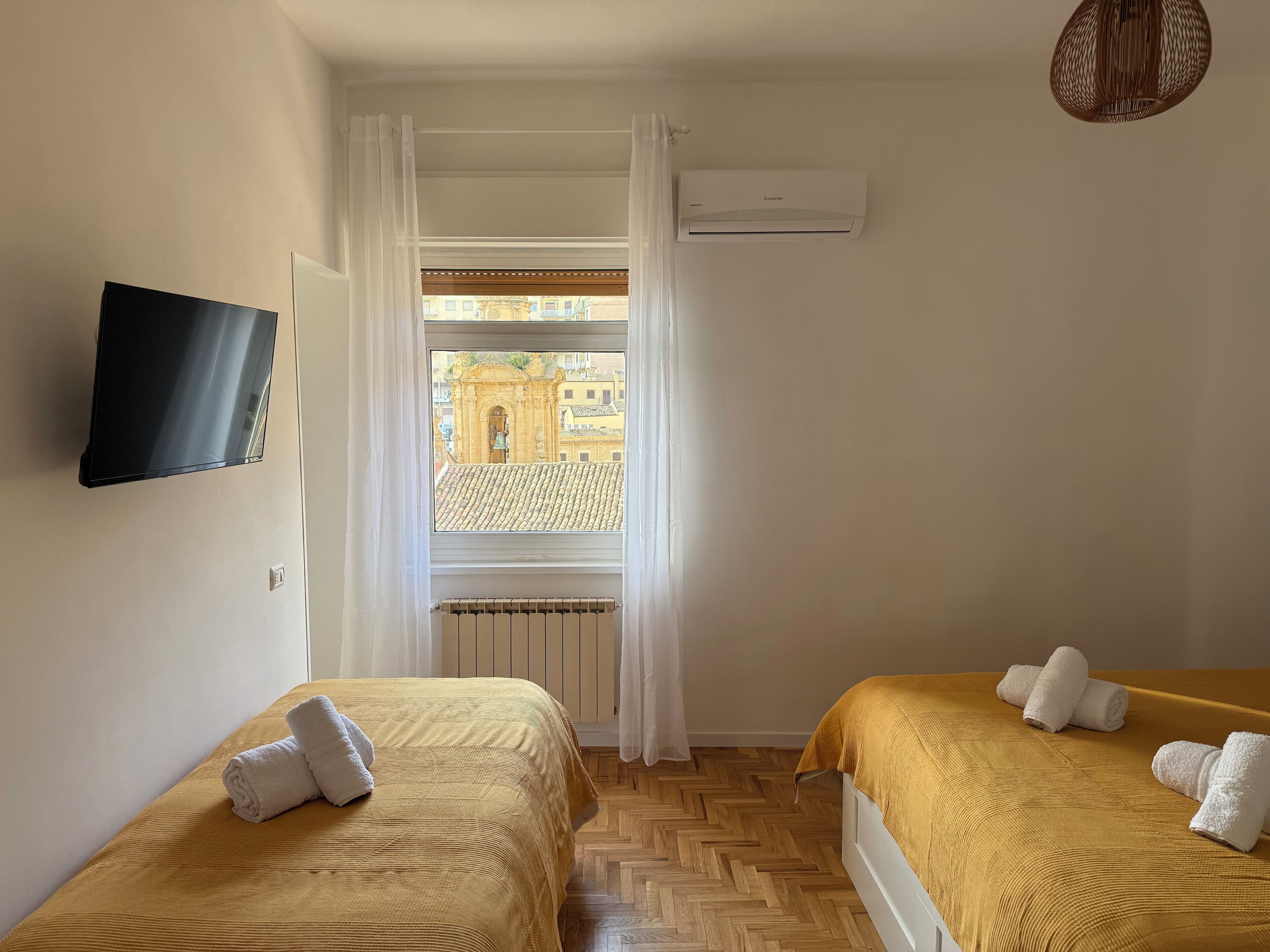 A hotel room with two beds, a TV, an air conditioner, and a window with a view of buildings.