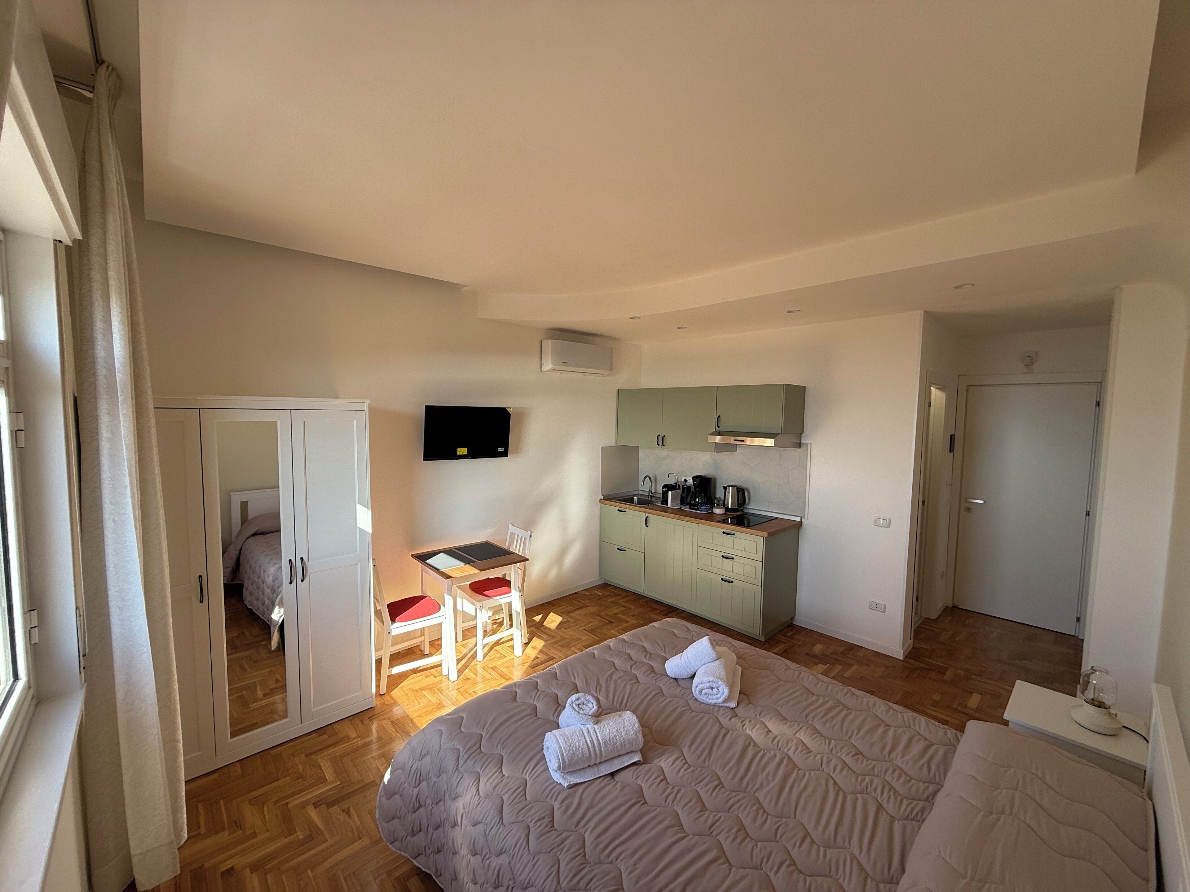 A compact hotel room with a bed, a small desk with a chair, a kitchenette, and a bathroom.