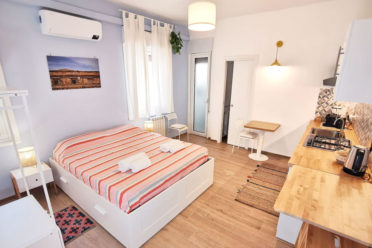 Quarto com cama, mesas de cabeceira, pequena escrivaninha, radiador e janela com cortinas.