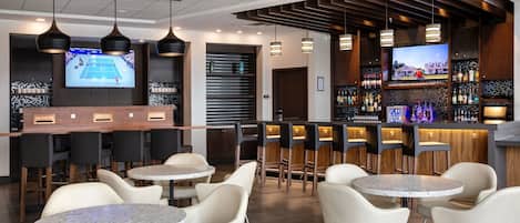 Un bar moderno con diversas opciones de asientos, incluyendo mesas redondas y taburetes de bar. Hay televisores montados en las paredes, y se exhiben una variedad de botellas y vasos en estanterías.