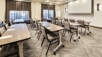 A conference room with multiple tables and chairs, a projector screen, and a large window with curtains.