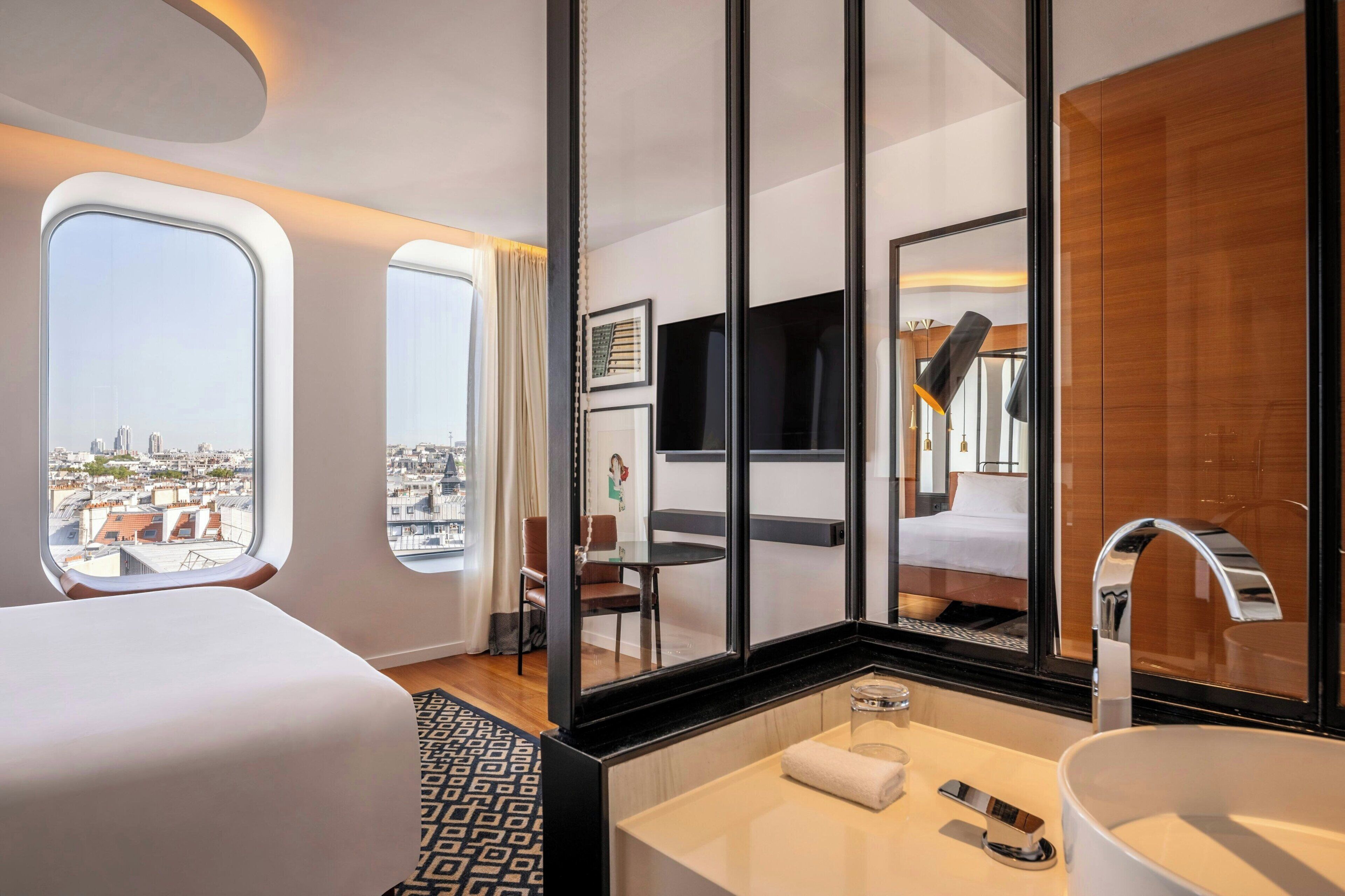 Ett modernt hotellrum med en stor säng, ett badrum med handfat och spegel samt stora fönster med utsikt över staden.