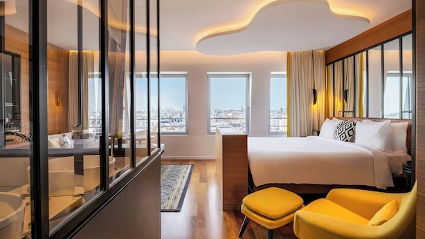 A modern hotel room with a large bed, a seating area with yellow chairs, a glass partition, and large windows offering a city view. - Renaissance Paris Republique Hotel (Paris)