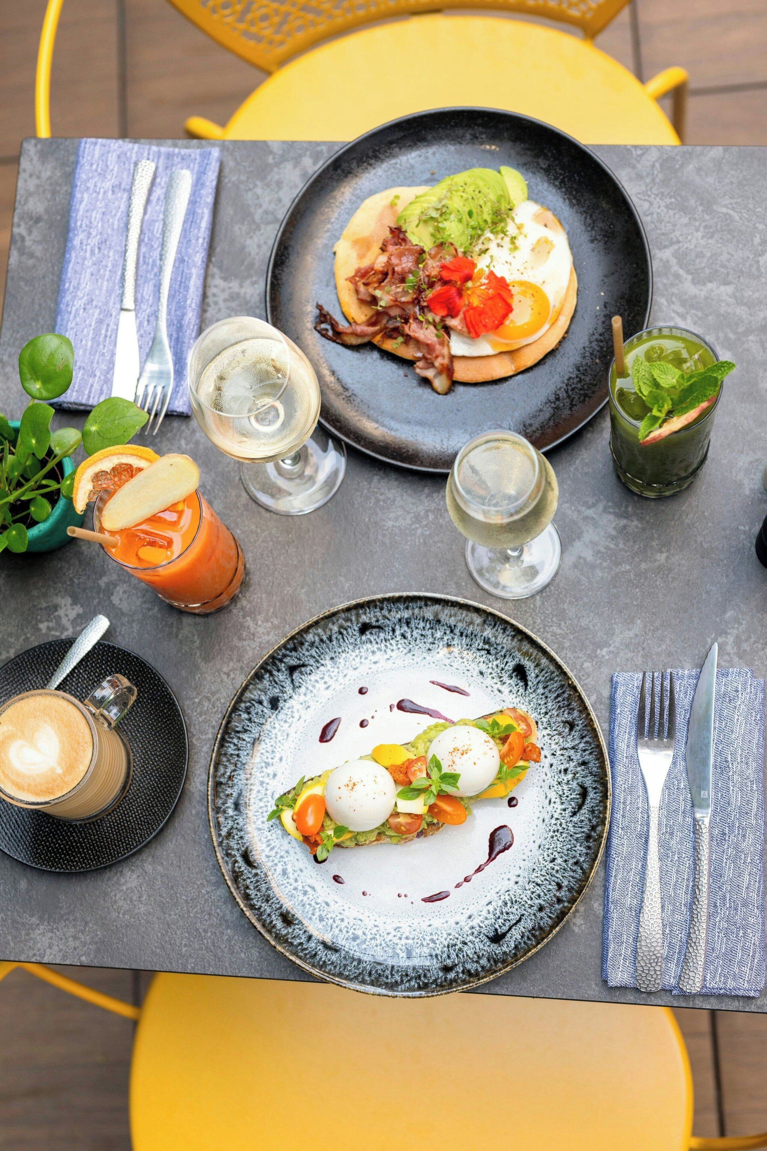 A table set with two plates of food, two glasses of white wine, a cocktail with a lime garnish, and a coffee drink.