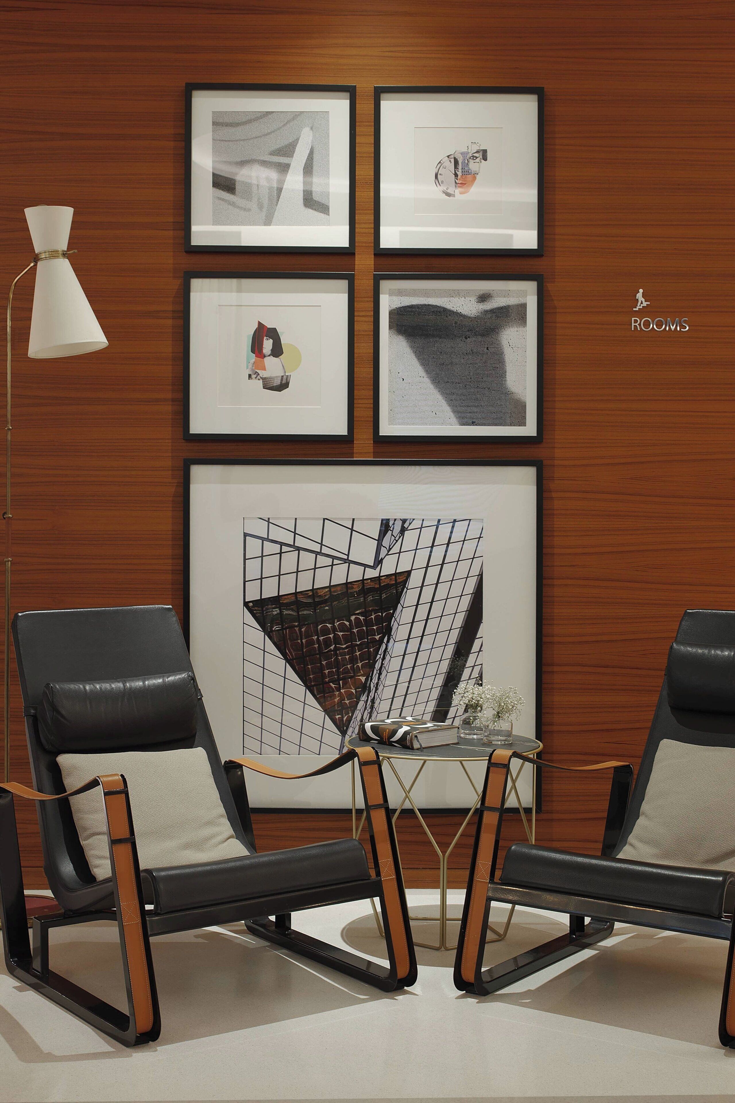 A modern room with two black leather chairs, a white floor lamp, and a table with a vase of flowers. There are four framed artworks on the wall.