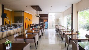 A spacious restaurant with multiple dining tables and chairs, a buffet area on the left, and large windows on the right.