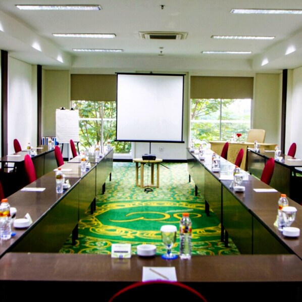 Un salón de conferencias con una larga mesa, sillas rojas, una pantalla de proyector y vistas al verdor exterior.