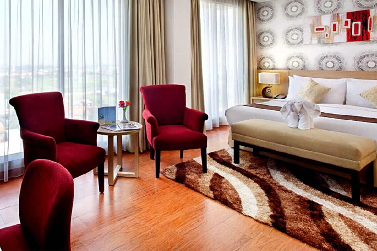 Quarto de hotel com uma cama grande, duas poltronas vermelhas, uma mesinha com um vaso e vista para a cidade através de grandes janelas.