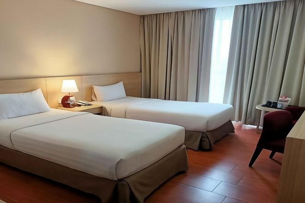A hotel room with two beds, a nightstand with a lamp, a small table with a chair, and a view of the outside through large curtains.