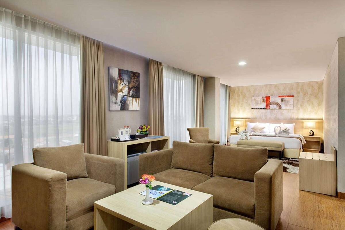 Quarto de hotel com sofá, mesinha com um vaso de flores, cama com edredom branco e uma obra de arte na parede.