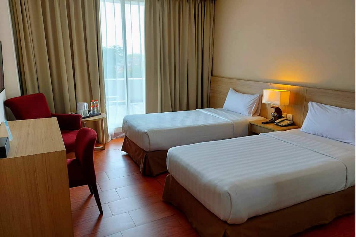 Quarto de hotel com duas camas, uma escrivaninha, uma cadeira e uma janela com cortinas.