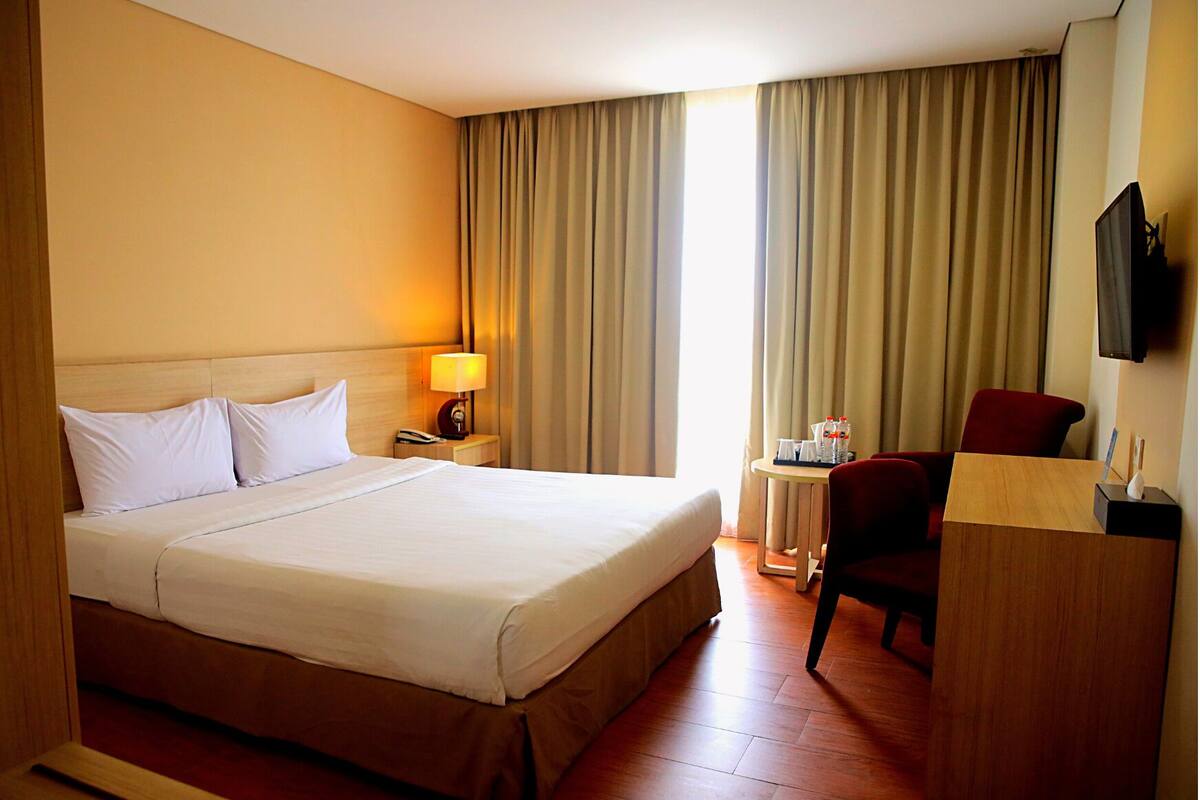 Quarto de hotel com uma cama grande, uma escrivaninha com cadeira, uma TV e uma mesinha de cabeceira com abajur.