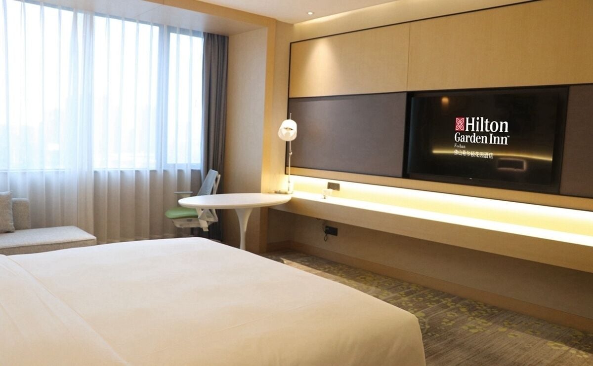 Quarto de hotel com uma cama grande, uma mesa de cabeceira, uma TV na parede e uma janela com cortinas.