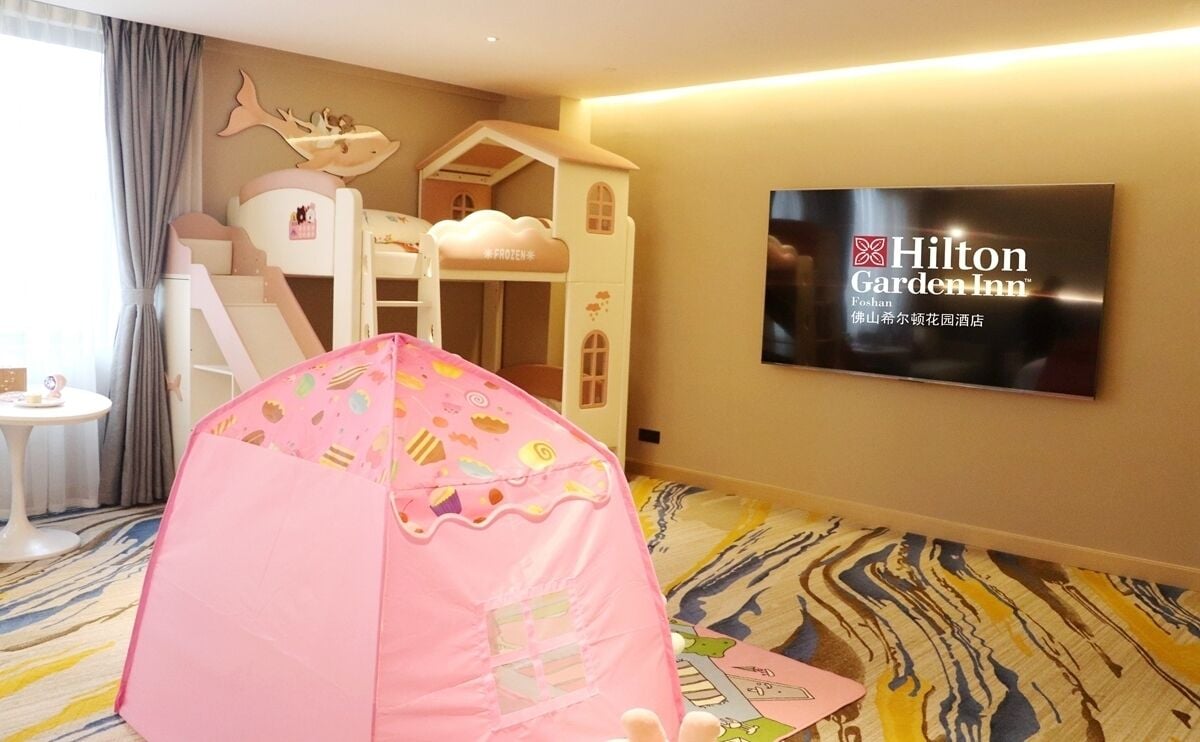 A hotel room with a bunk bed, a pink tent, a television displaying the Hilton Garden Inn logo, and a patterned carpet.
