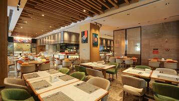 A modern dining area with multiple tables and chairs, a kitchen in the background, and a wooden ceiling.