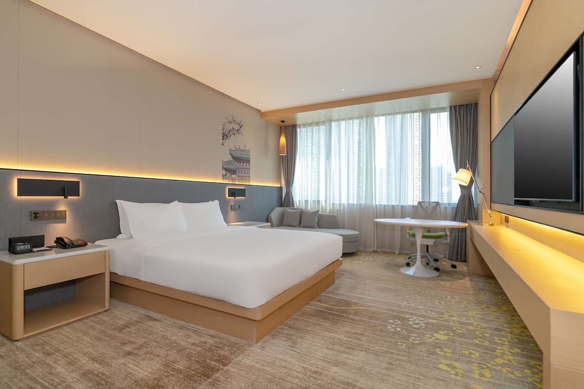 A modern hotel room with a large bed, bedside tables, a desk, a chair, a TV, and a large window with curtains.