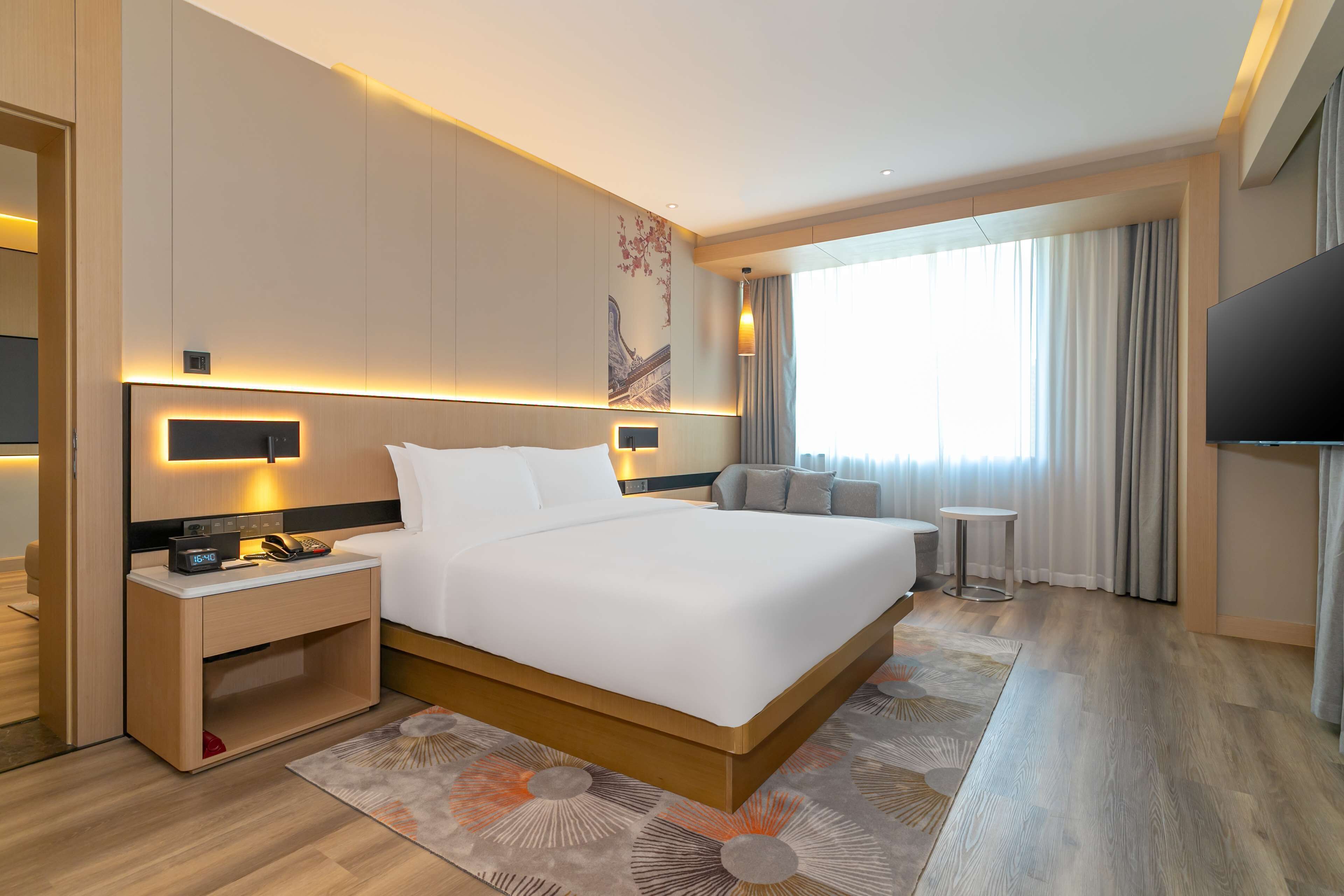A modern hotel room with a large bed, bedside tables, a flat-screen TV, a small side table, and a large window with curtains.