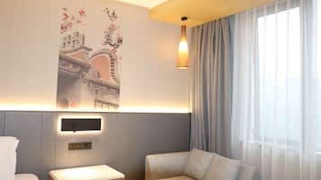 A hotel room with a bed, a bedside table, a sofa, a wall-mounted light, and a large window with curtains.