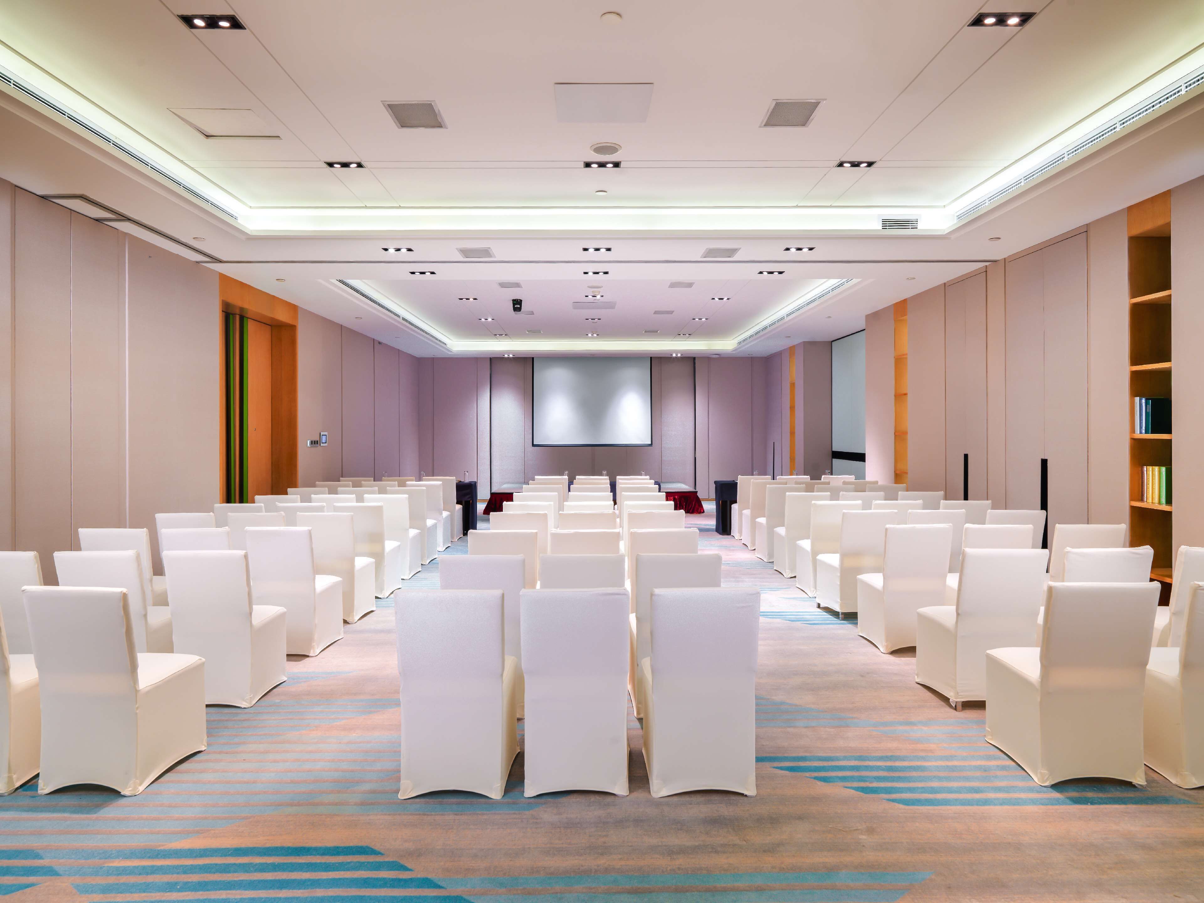 A conference room with rows of white chairs, a large screen, and a projector.