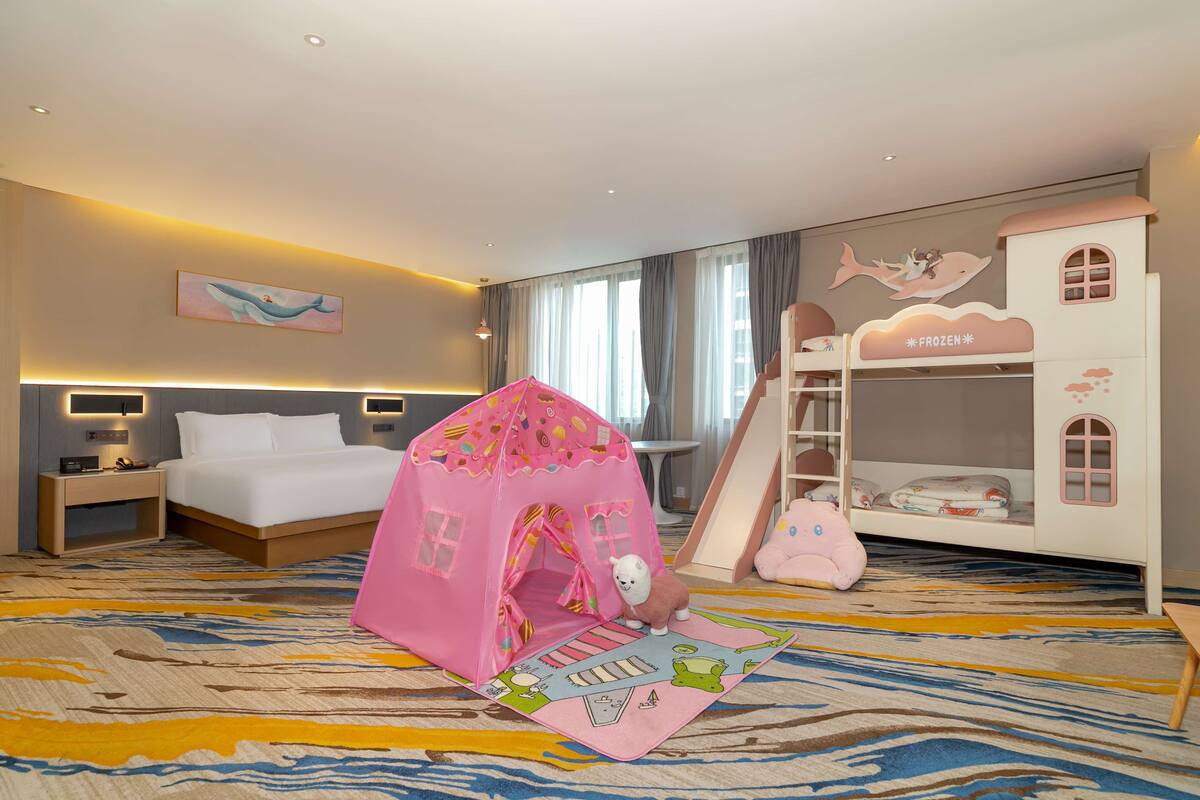 A hotel room with a bed, a pink playhouse, a bunk bed, and a large window.