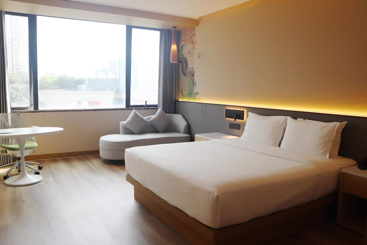 A hotel room with a large bed, a small table, a chair, and a view of the city.