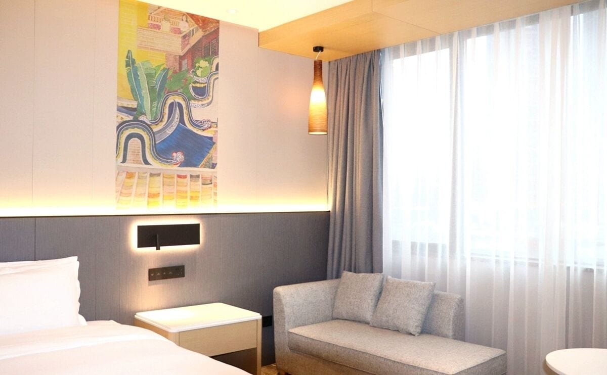 Ein Hotelzimmer mit Bett, Schlafsofa, Nachttisch, Wandlampe und einem großen Fenster mit Vorhängen.