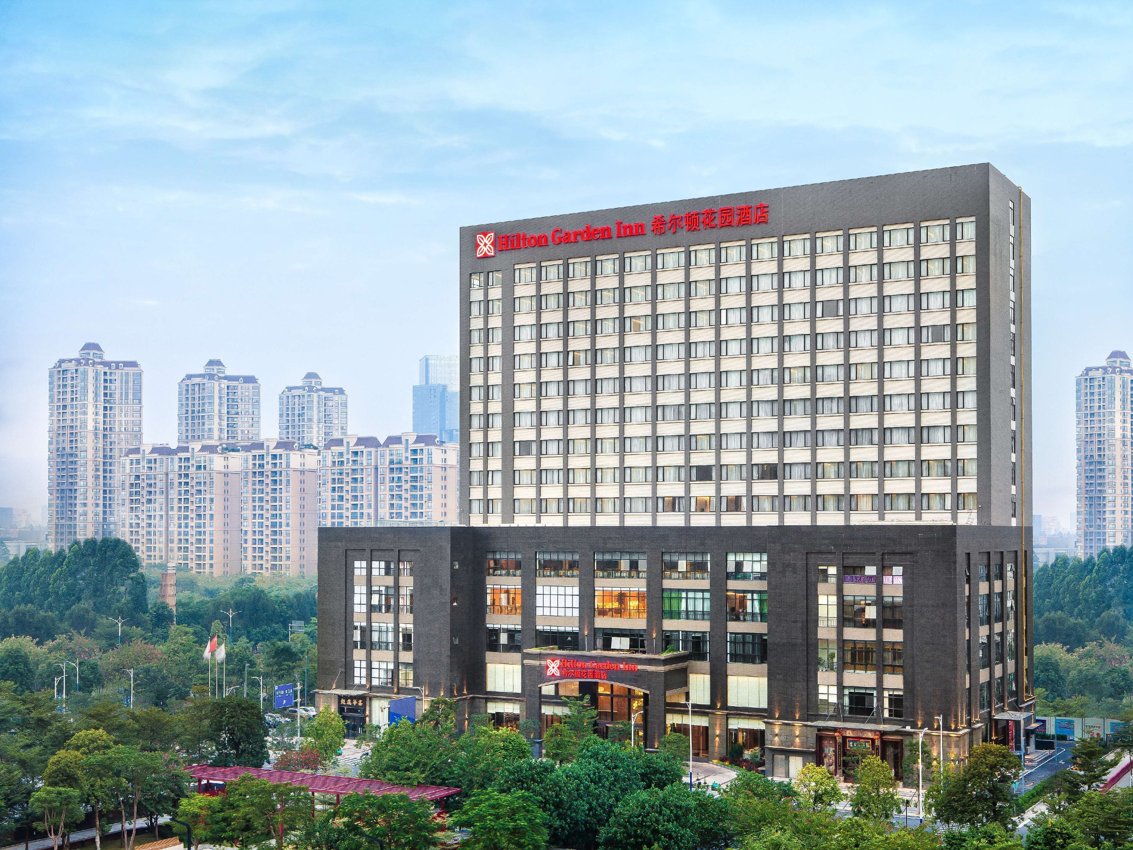 Un moderno edificio de hotel con un llamativo letrero Hilton Garden Inn. Presenta una estructura de varios pisos con fachada de vidrio y ladrillo, varios locales comerciales a nivel de calle, y está rodeado de vegetación y otros edificios altos.