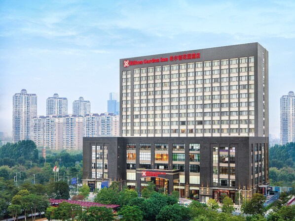 A modern hotel building with a prominent Hilton Garden Inn sign. It features a multi-story structure with a mix of glass and brick facade, several ground-level commercial spaces, and is surrounded by greenery and other high-rise buildings. - Hilton Garden Inn Foshan (Foshan)