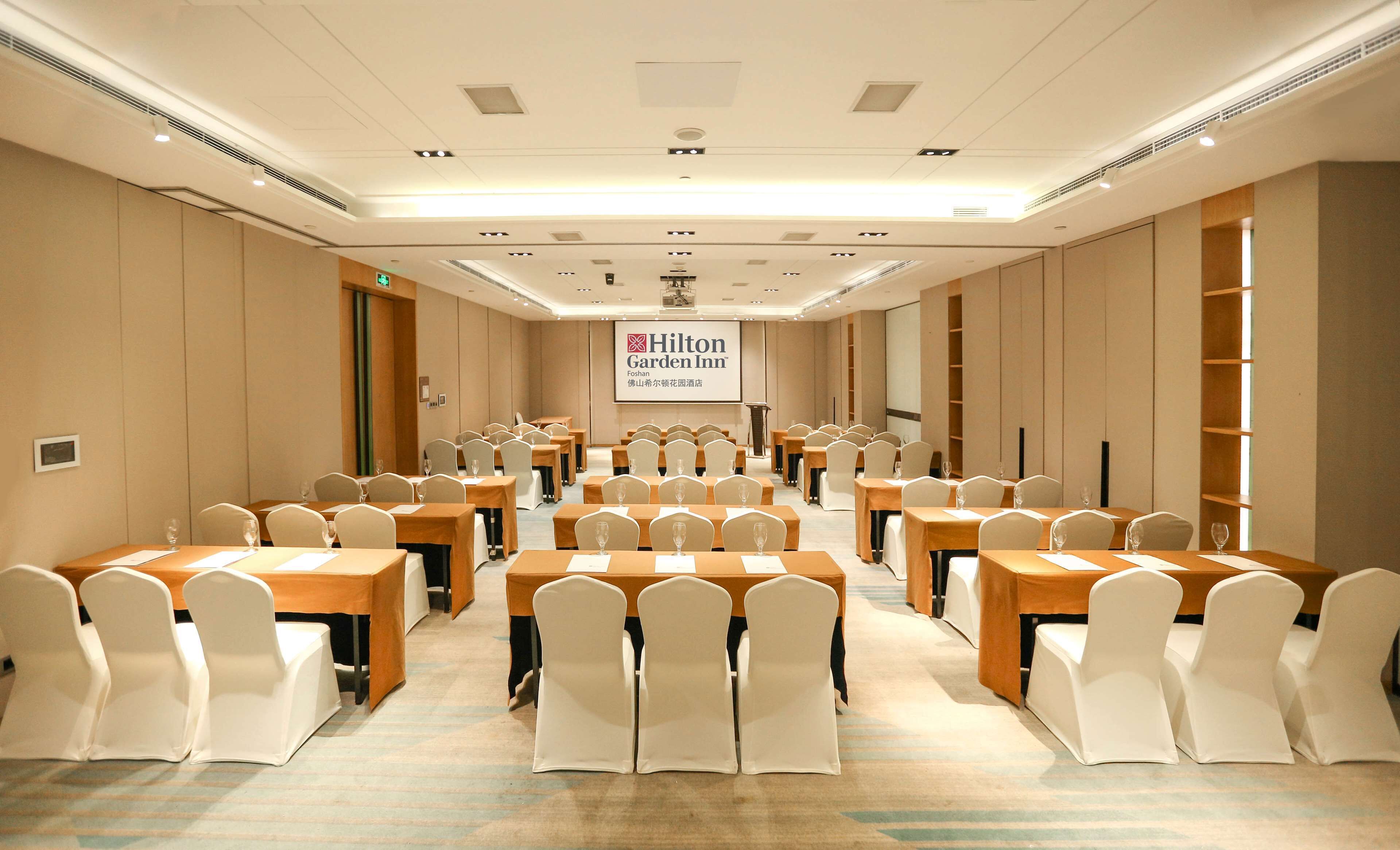 A conference room with multiple tables and chairs, a large screen displaying the Hilton Garden Inn logo, and a carpeted floor.