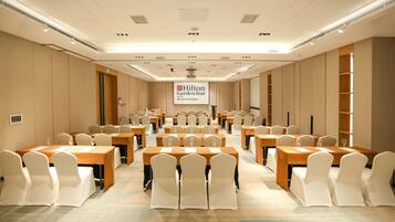 A conference room with multiple tables and chairs, a large screen displaying the Hilton Garden Inn logo, and a carpeted floor.