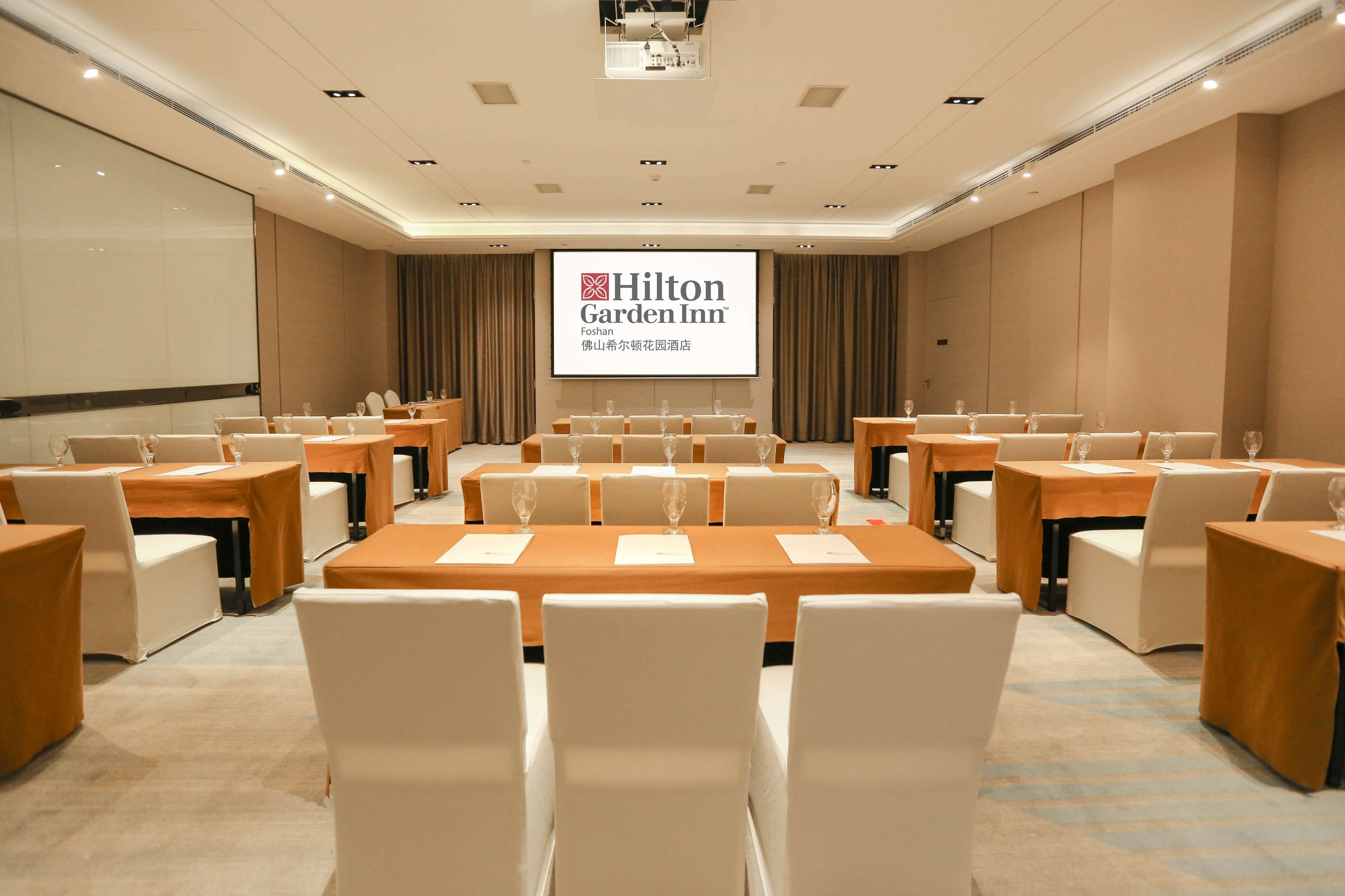 A conference room at Hilton Garden Inn with multiple tables and chairs, a large screen displaying the hotel name, and a carpeted floor.
