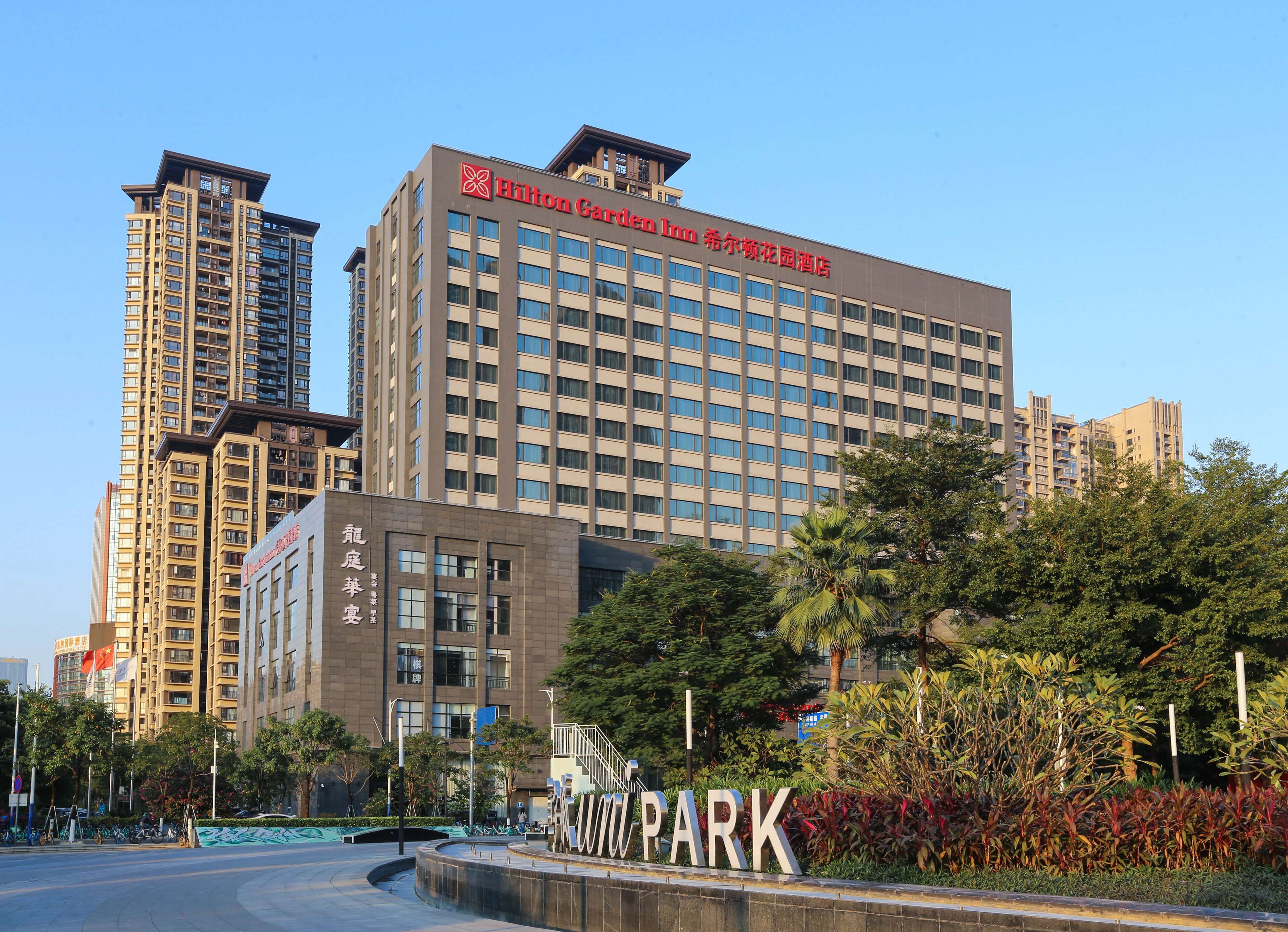 A modern hotel building with a prominent Hilton Garden Inn sign, surrounded by lush greenery and a well-maintained park area.