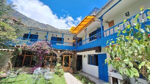 Un edificio de dos pisos con paredes exteriores azules y blancas, un balcón con toldo amarillo, y un jardín con plantas en macetas y mobiliario exterior.
