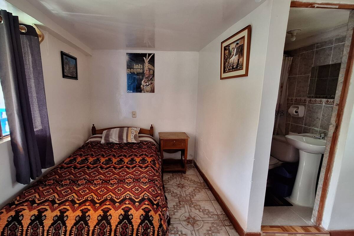 A small, compact room with a bed, a nightstand, a framed picture on the wall, and a bathroom visible through an open door.
