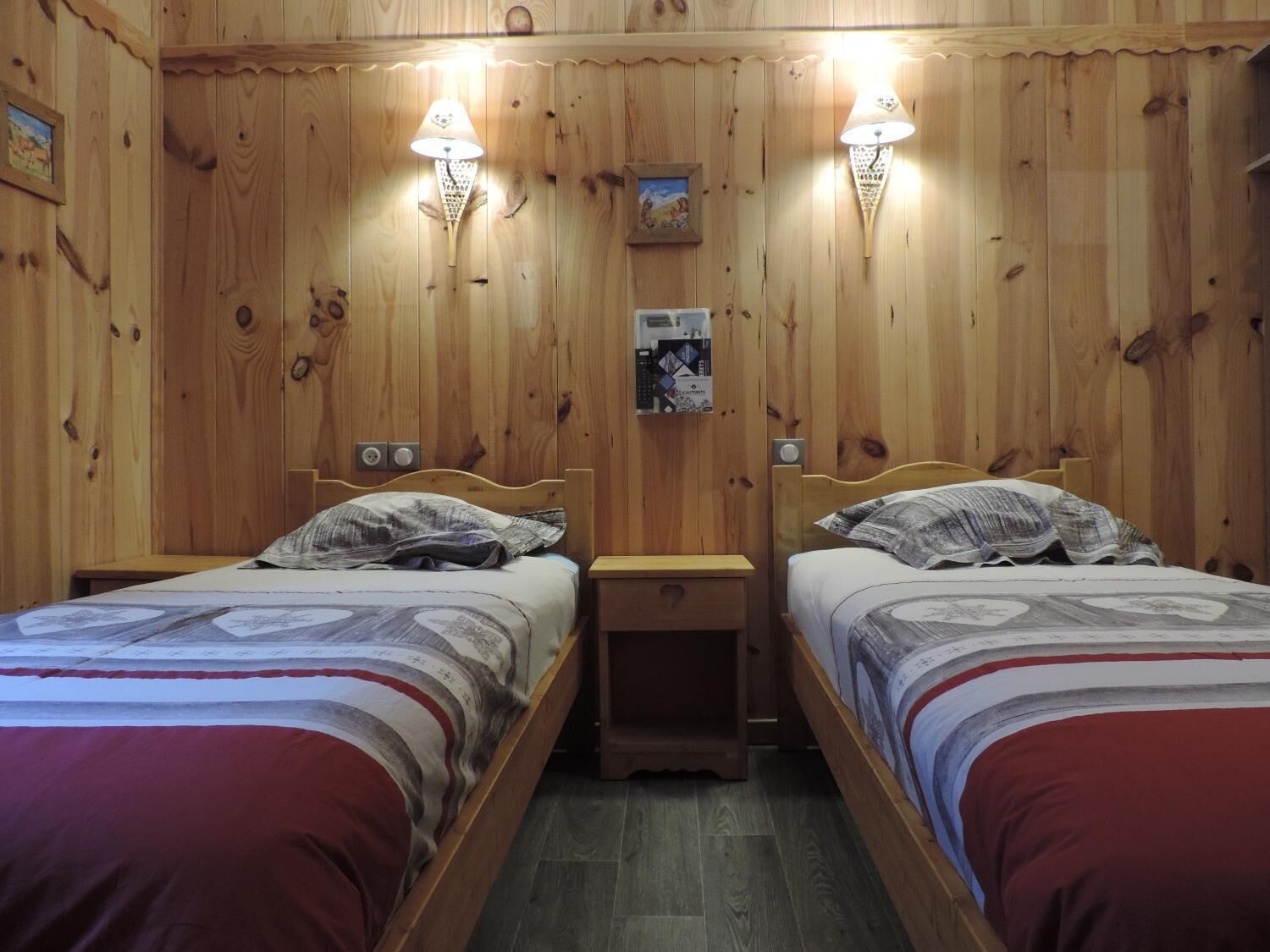Une chambre avec deux lits en bois, chacun muni d’une table de chevet et d’une lampe fixée au mur. Des tableaux encadrés ornent le mur, et une petite étagère contient des livres.