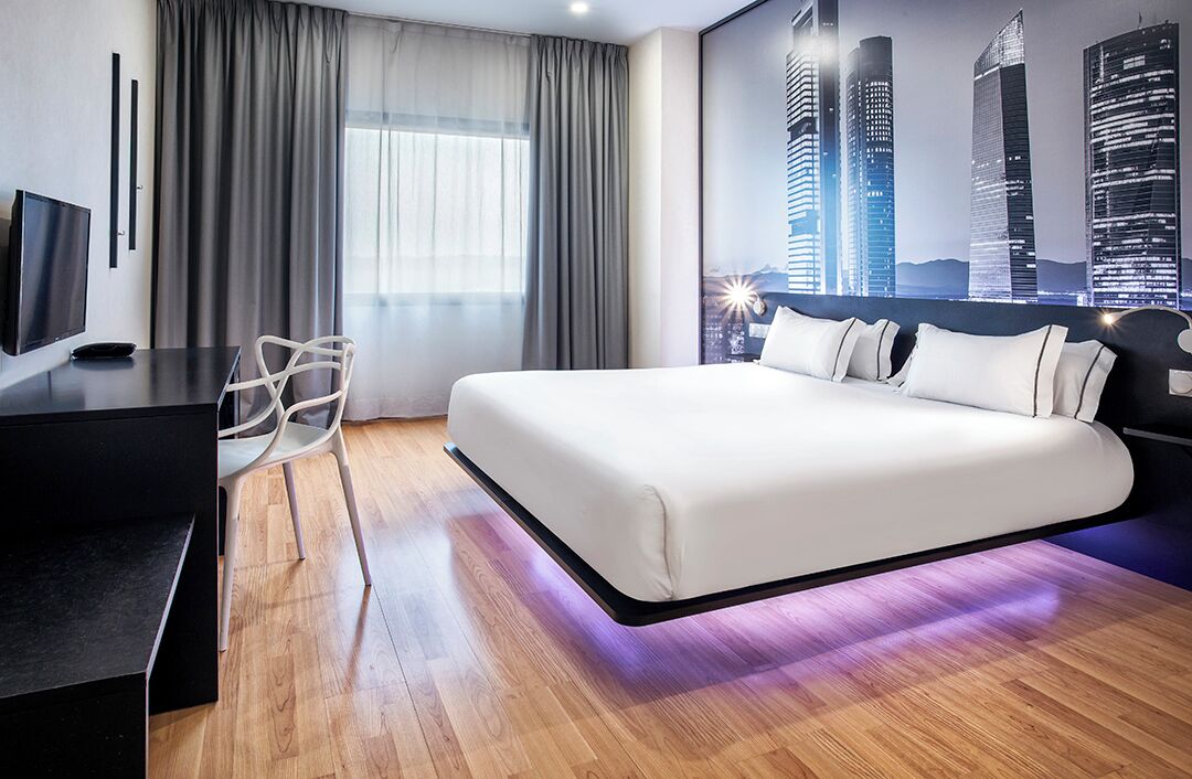 A modern hotel room with a large bed, a desk, a chair, and a cityscape view.