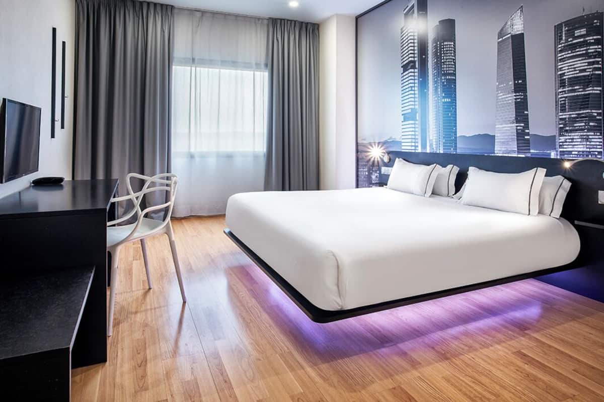 A modern hotel room with a large bed, a desk, a chair, and a cityscape view.