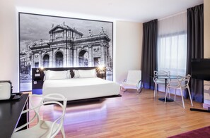 Ein modernes Hotelzimmer mit einem großen Bett, einem Schreibtisch mit Stuhl, einem kleinen Tisch mit Sitzgelegenheiten und einem großen Fenster mit Vorhängen.