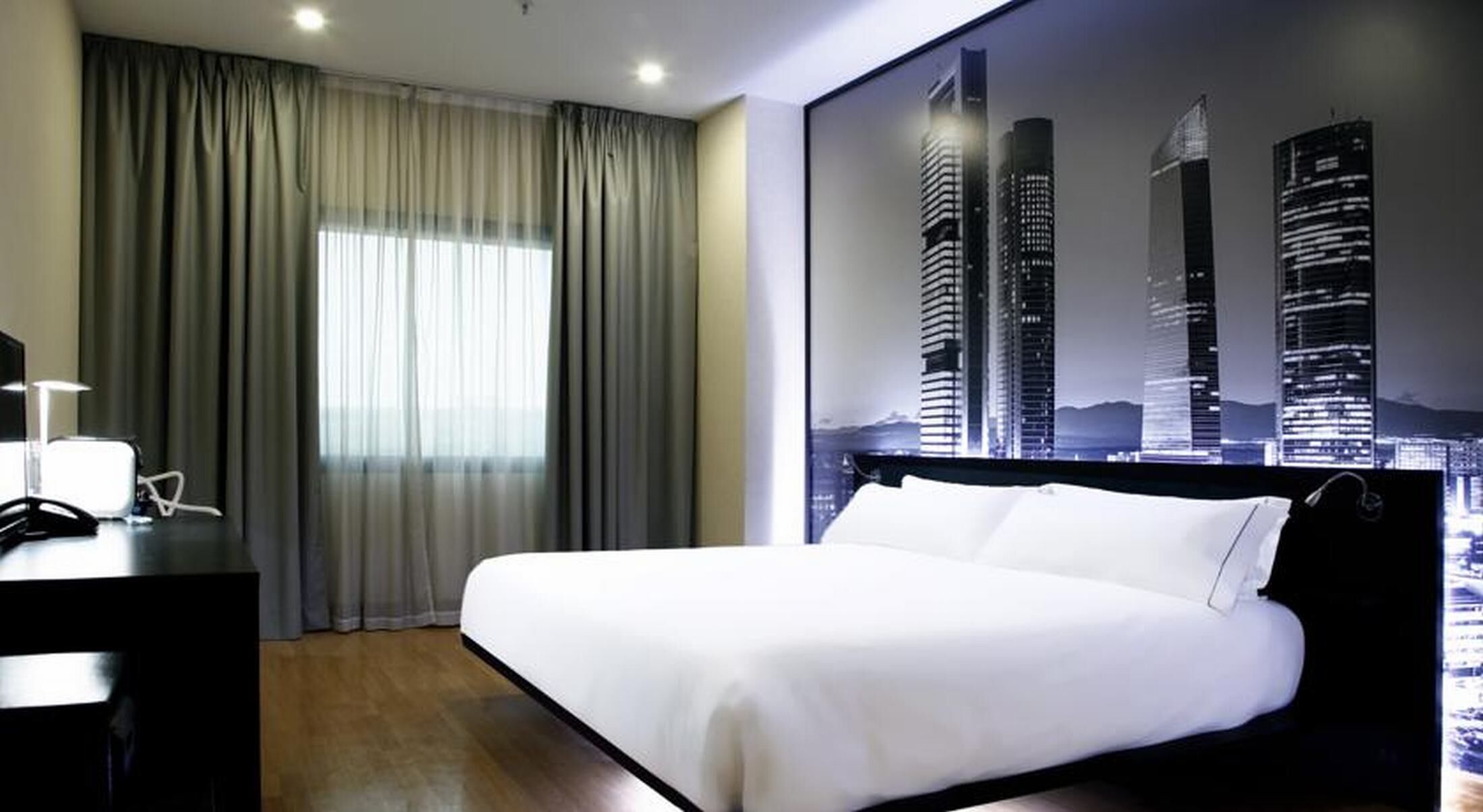 A hotel room with a bed, curtains, a desk with a lamp, and a cityscape view.