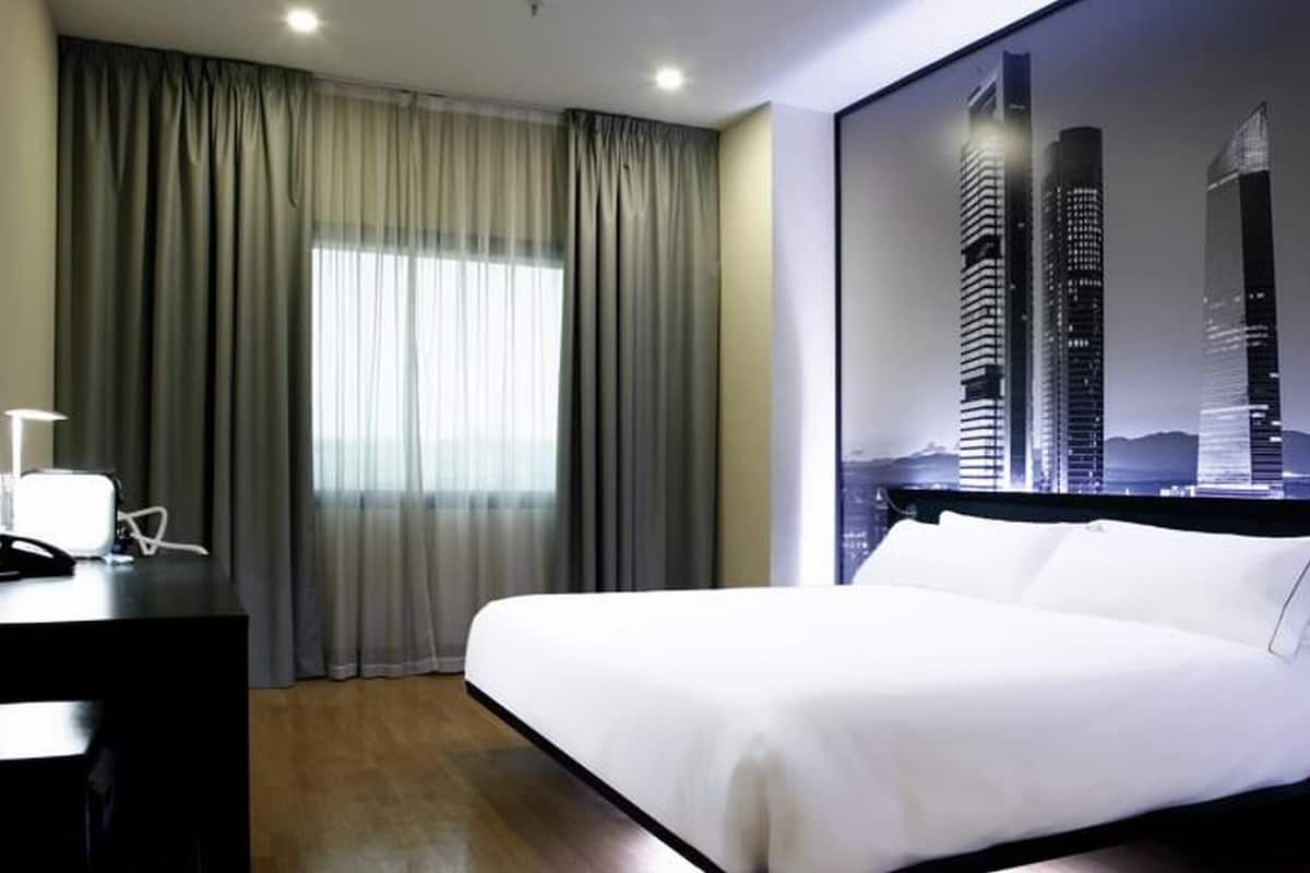 A hotel room with a bed, curtains, a desk with a lamp, and a cityscape view.