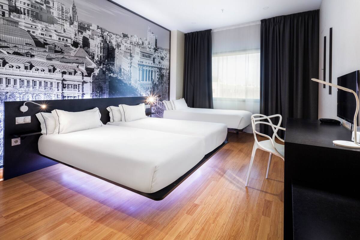 Ein modernes Hotelzimmer mit einem großen Bett, einem kleineren Bett, einem Schreibtisch mit einem Flachbildfernseher und einer Stadtlandschaftsmalerei an der Wand.