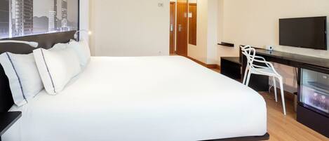 A hotel room with a large bed, a desk with a chair, a television, and a refrigerator.