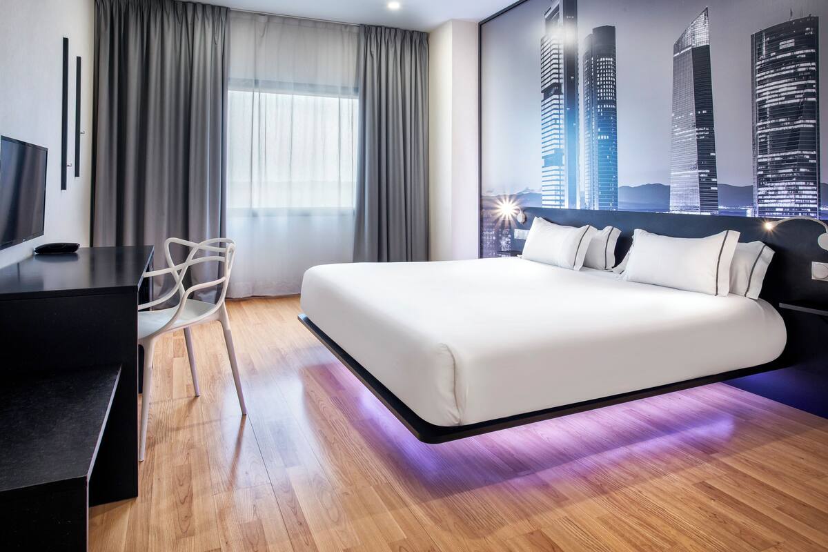 Ein modernes Hotelzimmer mit einem großen Bett, einem Schreibtisch und einem Flachbildfernseher, sowie einem Wandbild mit Stadtansicht.