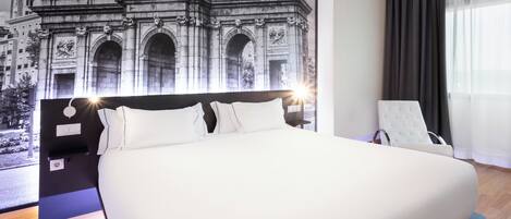 Una habitación de hotel moderna con una cama grande, un cabecero con un mural de un paisaje urbano, un sillón blanco y un ventanal con cortinas translúcidas.