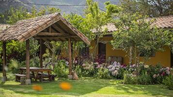 Un pavillon rustique en bois, doté d’une table de pique-nique, est entouré de parterres luxuriants où poussent différentes variétés de fleurs, avec une chaîne de montagnes en toile de fond.