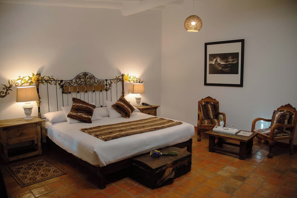 A neatly arranged bedroom with a large bed, two bedside tables with lamps, a wooden bench, two chairs, and a framed picture on the wall.