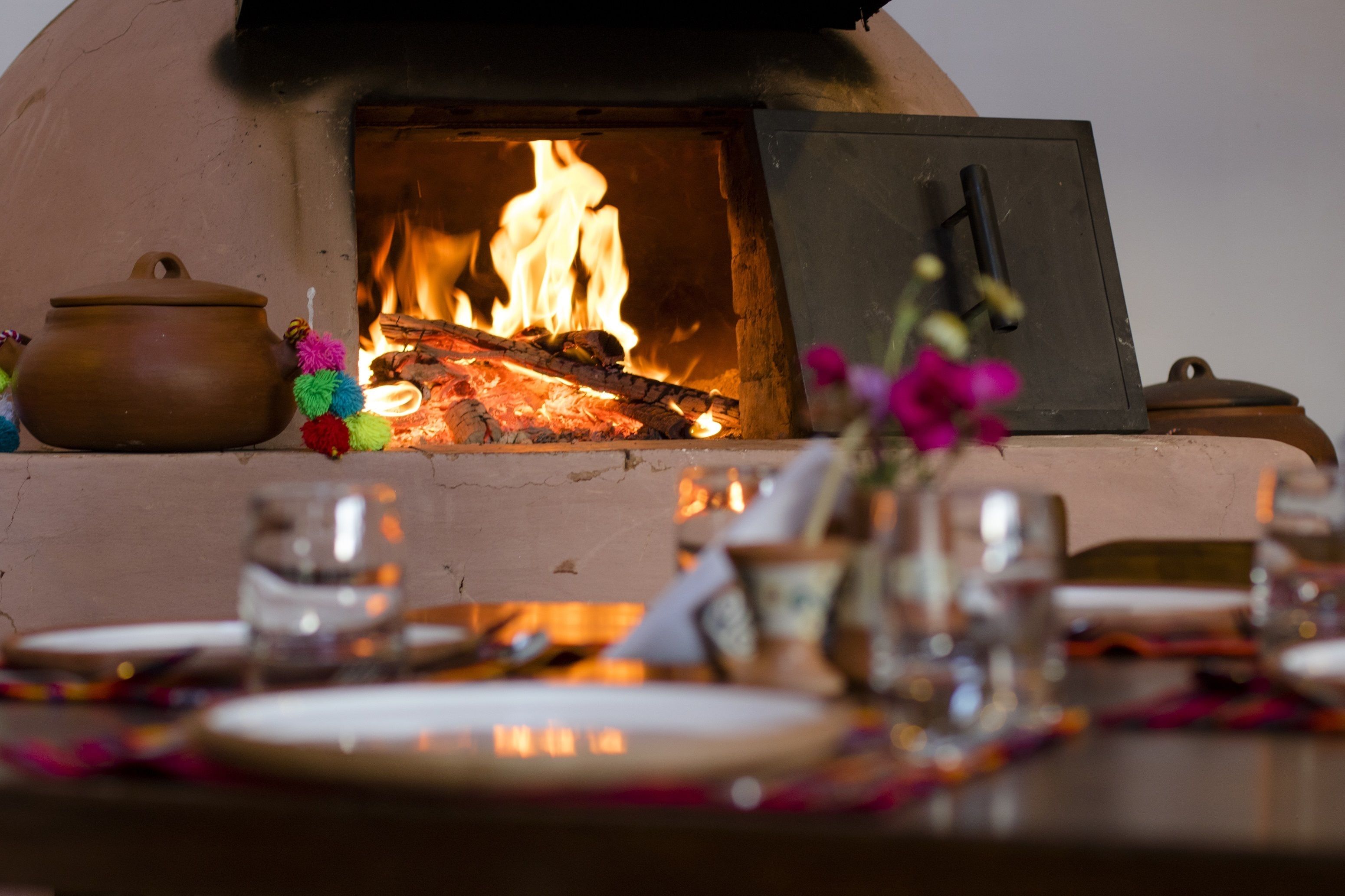 A traditional wood-fired oven with a pot on the left, a glass on the right, and a table set with plates and a vase of flowers.