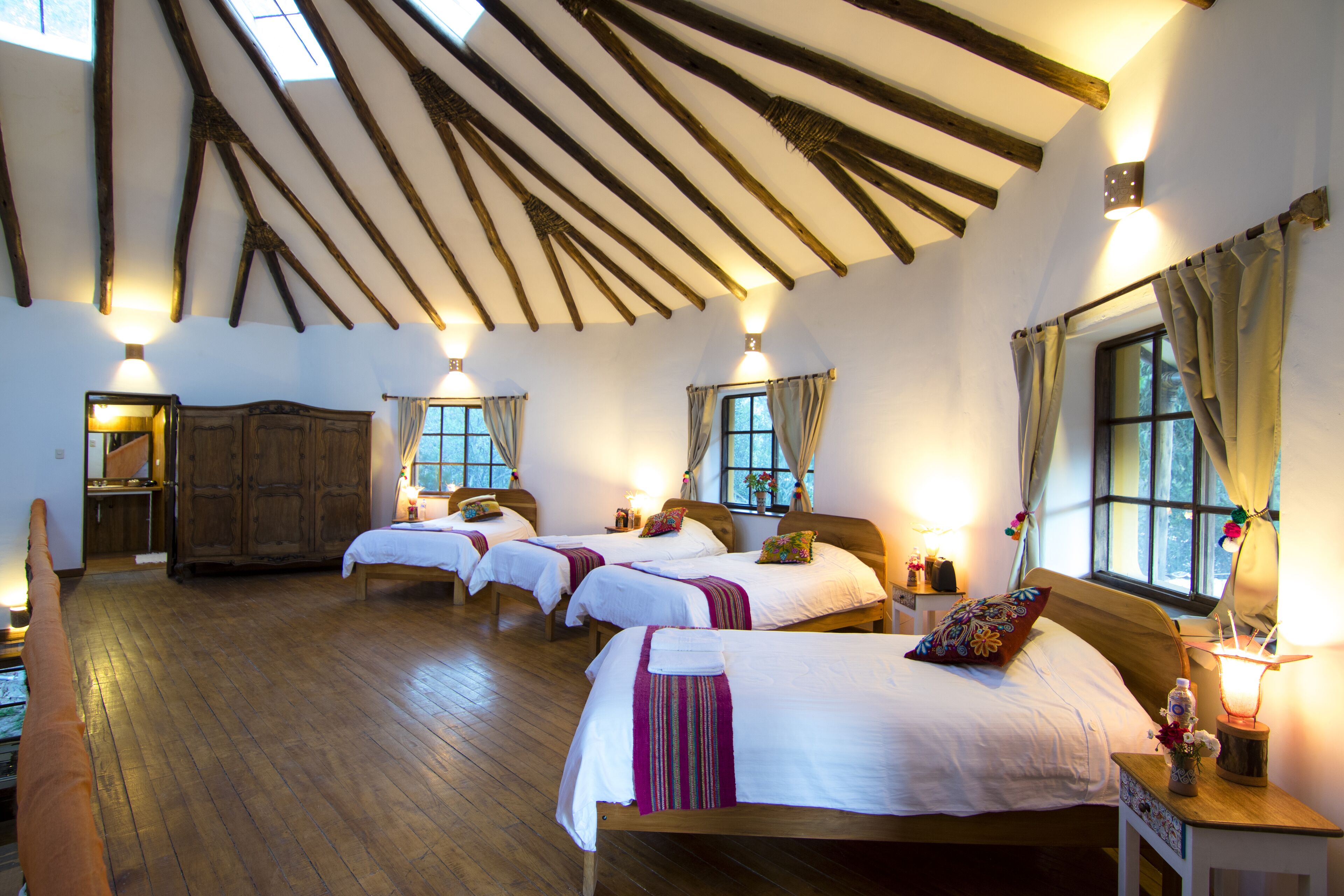 Quarto com quatro camas, vigas de madeira no teto e janelas com cortinas.